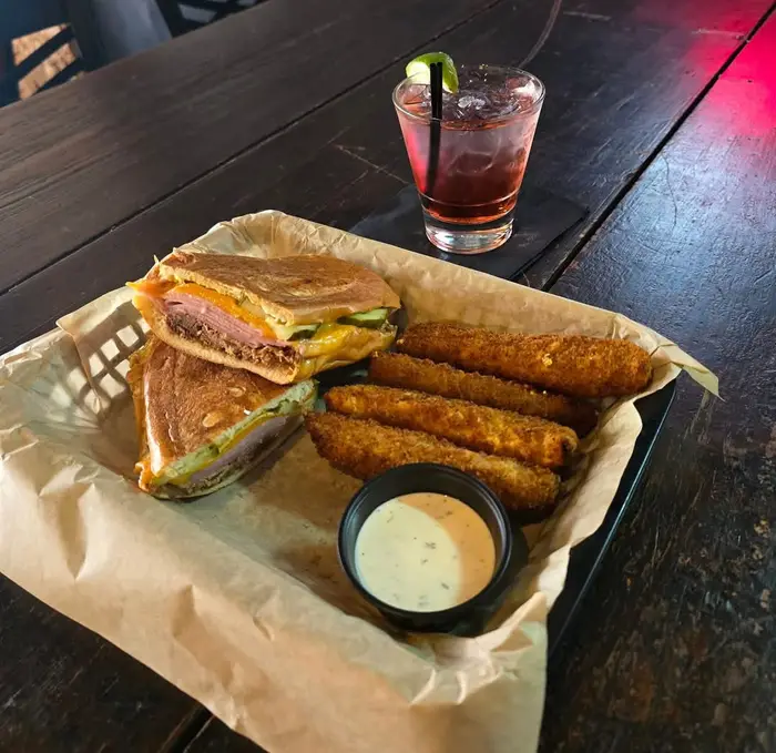 The OTP Cuban cut in half in a paper lined basket with breaded pickle spears and a cocktail on the table
